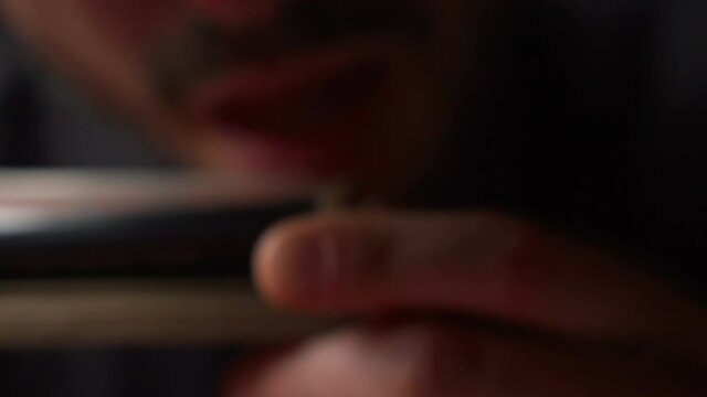 A man holds a cigarette rolling machine in his hands and raises it to his face and wets the edge of the paper with his tongue to seal the joint twisted from marijuana