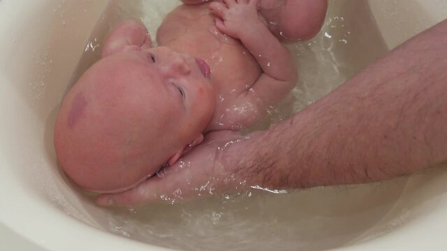 Father holding baby in bath bathing and washing, 1 month old infant in plastic tub in bathroom at home. High quality 4k footage