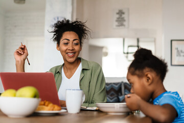 Black smiling woman working while having breakfast with her daughter