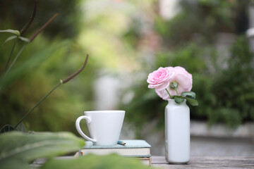 Fototapeta na wymiar white coffee cup with pink rose in ceramic vase and notebooks on weathered wooden table at outdoor