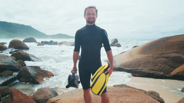 Surf photographer. Portrait of caucasian surfing photographer standing on the rocky coast with fins and underwater housing in hands