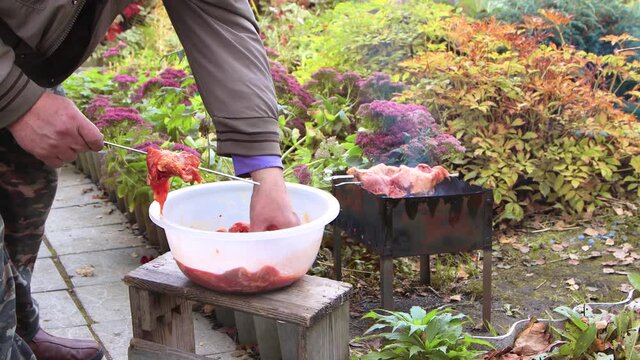Male hands string pieces of meat on a skewer in a garden with flowers. The recipe for cooking barbecue on charcoal. Making food over an open fire. Outdoor picnic in the garden. Warm day, soft sunshine