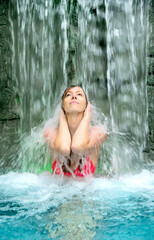 young sexy woman in pink bikini holds hands on head, enjoy the falling water of the waterfall in the spa pool, copy space
