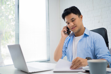 Young Asian businessman talking on the phone and looking at the notebook at his white desk.
