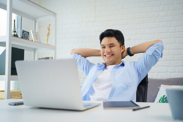 Fototapeta premium Young Asian man is smiling and reading the screen on laptop while relaxing and working at home.