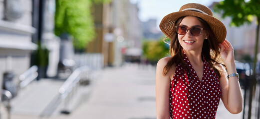 Young smiling girl (female) in sunglasses straw hat and red dress with polka dots walks in the...