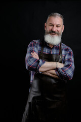Portrait of handsome mature bearded man dressed leather apron isolated on black background, caucasian workman with beard smiling