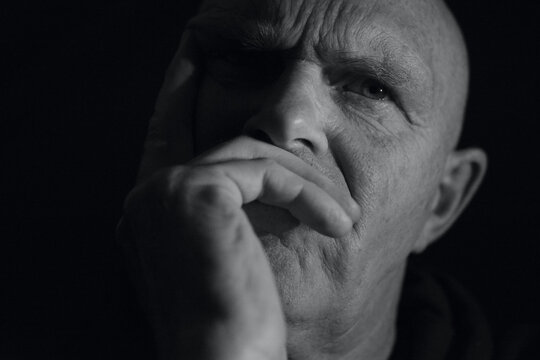 Close up portrait of a mature make with hand over his moth looking puzzled and frowning. Black and white photography with a head shot against a black background