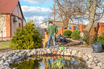 Mature caucasian man with his dog cleans a garden pond from water plants and falling leaves. Spring seasonal pond care after winter.