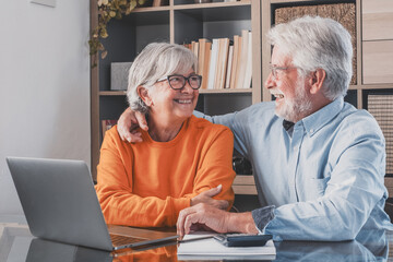 Happy elderly 60s couple sit rest on couch at home pay household expenses online on computer, smiling mature 50s husband and wife clients hold documents make payment on internet banking laughing
