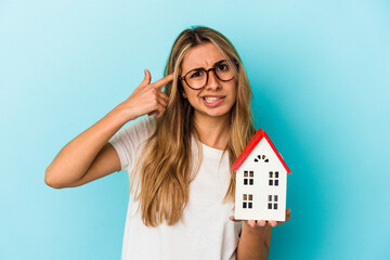 Young caucasian woman holding a house model isolated on blue background showing a disappointment...