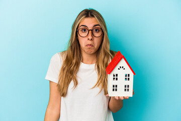 Young caucasian woman holding a house model isolated on blue background shrugs shoulders and open...