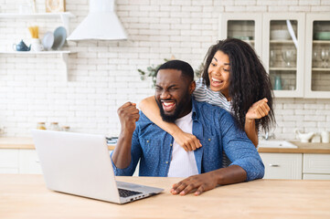 Biracial woman hugs african american man from behind at laptop, rejoices at her husband's success, cheerful guy got a promotion at work, won the lottery, bought a vacation ticket, sitting at laptop