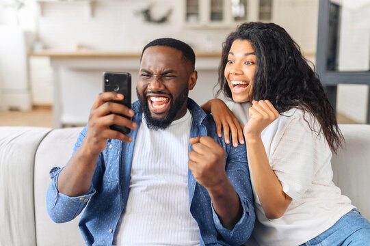 Black overjoyed couple resting on couch holding smart phone, celebrating on-line lottery win, bid betting victory moment, unbelievable opportunity or invitation, internet sale, getting prize concept