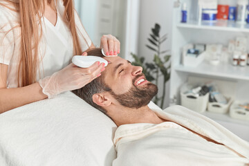 Obraz na płótnie Canvas Bearded man smiling in professional beauty spa salon during ultrasonic facial cleansing procedure