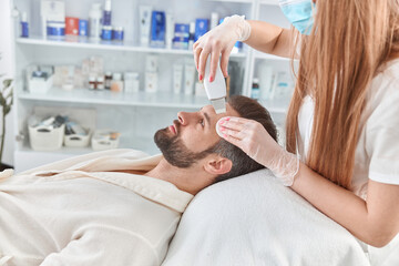 Obraz na płótnie Canvas Bearded man smiling in professional beauty spa salon during ultrasonic facial cleansing procedure