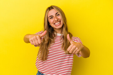 Young blonde caucasian woman isolated on yellow background raising both thumbs up, smiling and confident.