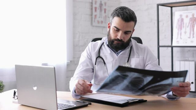 Young male doctor is looking at chest x-ray while sitting in medical office. Specialist examining x-ray picture and holding it in his hands, taking notes in medical form during working day in hospital