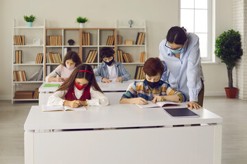 Coronavirus safety measures in class. Students and teacher wearing medical face masks during Covid-19 pandemic. Primary school children with mouths covered writing in notebooks in modern classroom