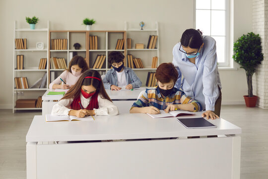 Coronavirus safety measures in class. Students and teacher wearing medical face masks during Covid-19 pandemic. Primary school children with mouths covered writing in notebooks in modern classroom