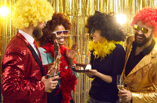 Group Of Happy Friends In Retro Outfits, Feather Boas And Afro Hair Wigs With Cake And Champagne Congratulating Young Black Woman At 70s Disco Music Nightclub Fancy Dress Fashion Themed Birthday Party