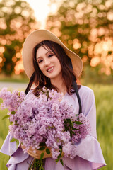 Stylish girl in violet dress and hat in field. Happy young woman on the sunset or sunrise in summer nature. Fashion and lifestyle concept.