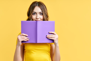 Young woman hiding behind an open book and looking to camera isolated over yellow background.