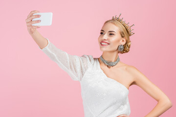 blonde woman in luxury crown taking selfie on smartphone isolated on pink