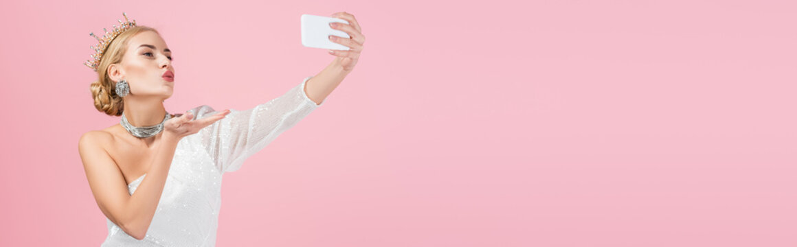 blonde woman in luxury crown taking selfie on smartphone and sending air kiss isolated on pink, banner