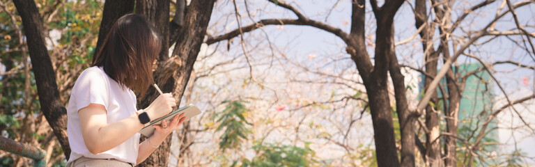 web banner asian woman solo traveler painting and diary record to her tablet with see pink cherry blossom blooming from springtime 