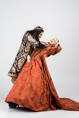 full length of cruel hispanic king in medieval clothing choking blonde queen in golden crown on white
