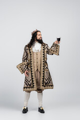 full length of hispanic king in medieval clothing and crown holding glass of red wine on white