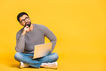 Portrait of a bearded young man in casual holding laptop computer while sitting on a floor isolated over yellow background. Copyspace for text. - Powered by Adobe