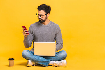 Young bearded man sitting on the floor with laptop and talking at phone. Isolated over yellow...