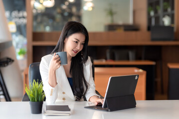 Fototapeta premium Charming Asian businesswoman holding a coffee mug working on a tablet at the office.