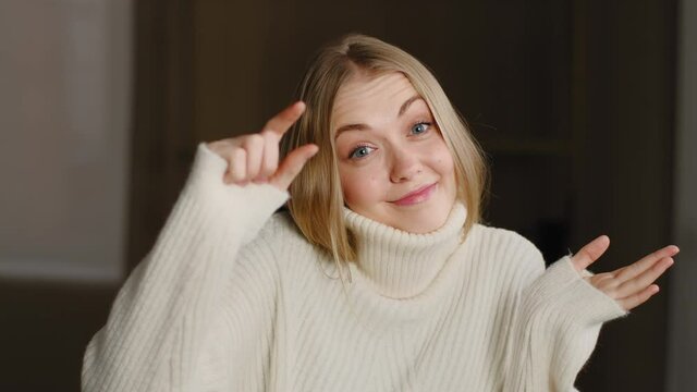 Young blonde model caucasian woman confused girl standing indoor, showing very tiny shapes small quantity minimum little size gesture with fingers, asking for piece, shrugging shoulders posing at home