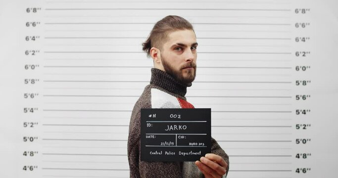 Mugshot of arrested bearded man turning head and looking to camera while standing aside . Crop view of handsome guy holding sign in police department. Concept of criminal.