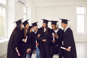 Group of students of different nationalities dressed in graduation gowns talk, smile and browse the album in the university classroom. Education, graduation and people concept.
