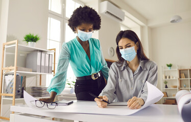 Safety during coronavirus pandemic. People in face masks work in office studio after end of lockdown and quarantine. Two creative designers and business women thinking and making poster or draft plan - Powered by Adobe