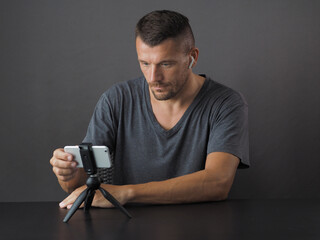 Man wearing gray t-shirt sitting at desk, using phone on tripod with wireless earphones. Concept online meeting.