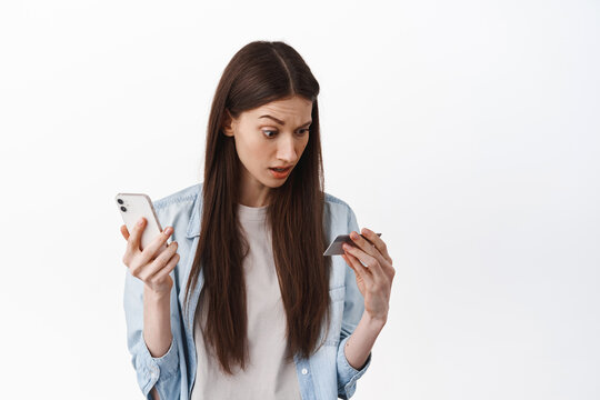 Online shopping. Woman holds smartphone and looks confused at credit card, have no money, checking her bank account in app, standing over white background