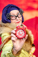 Fashionable grandmother in turban and glamorous glasses. Older woman shows alarm clock. Old woman in trend. Fashion for the elderly. Fashion for all ages. Trendy people. Elegant granny holding a clock