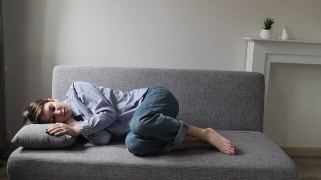 
A pensive sad girl with a short hair of nineteen years in a white shirt with blue stripes looks at the camera, lies on the couch. 