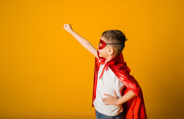 small boy in a suit with a red mask and cape stands sideways on a yellow background with space for text