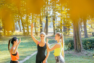 Fototapeta premium Group of three female athletes give each other high five after a good training session in fall park. Attractive sporty caucasian woman with two young fitness friends smiling outdoor.