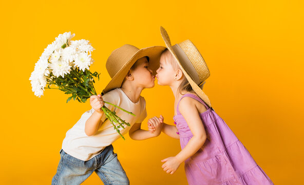 portrait of a little girl and a boy in straw hats kissing on a yellow background with space for text. A boy holds a bouquet of white flowers