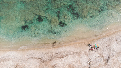 Aerial view from a drone, of the clear blue waters of a deserted beach, with three people lying on towels and a dog walking.