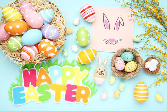 Text Happy Easter with colorful eggs in basket, mimosa flowers and picture of rabbit on blue background