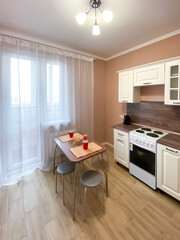 Interior of a beige kitchen with a white kitchen set and a stove, a table with utensils and a window with a balcony, covered with white tulle