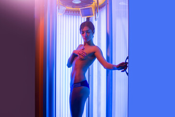 Young woman with sexy naked healthy body standing in solarium spa salon. Attractive young girl preparing for tanning in solarium. - obrazy, fototapety, plakaty